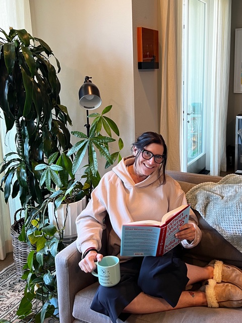 Andrea cozied up on the couch wearing a hoodie and Ugg slippers, holding a book and tea.